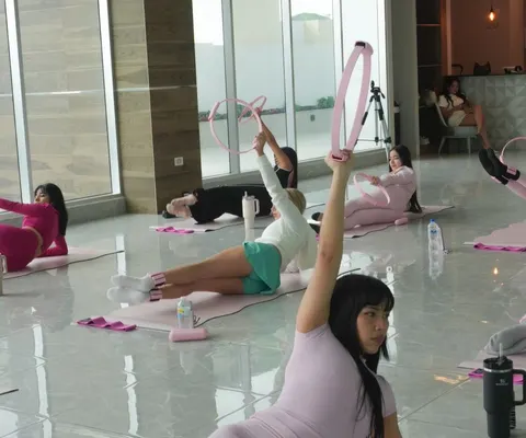 Una alumna sostiene un aro rosado de Pilates en alto mientras otras mujeres se mueven sobre mats rosados